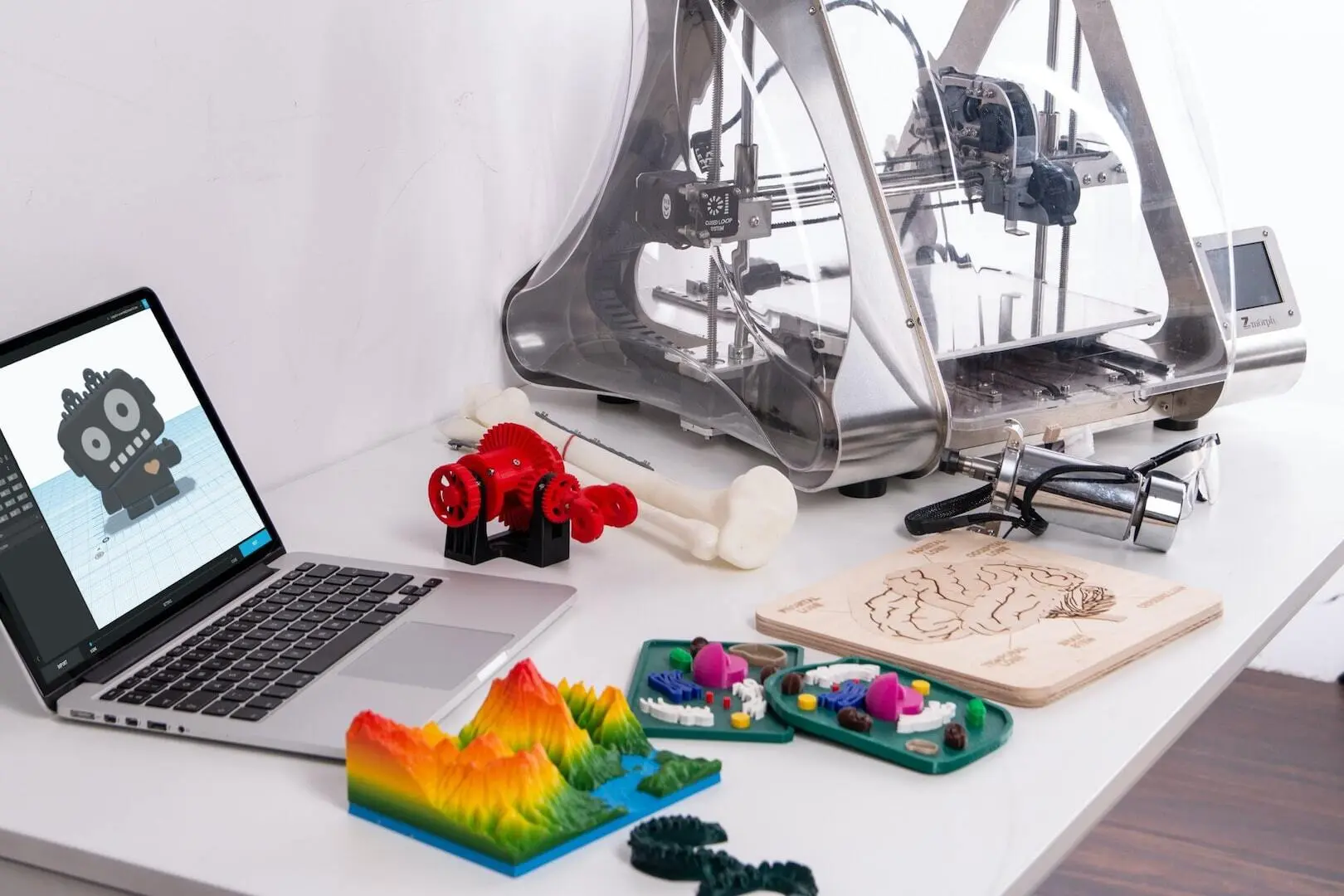 Laptop z modelem robota w programie 3D, drukarka 3D i wydruki na stole